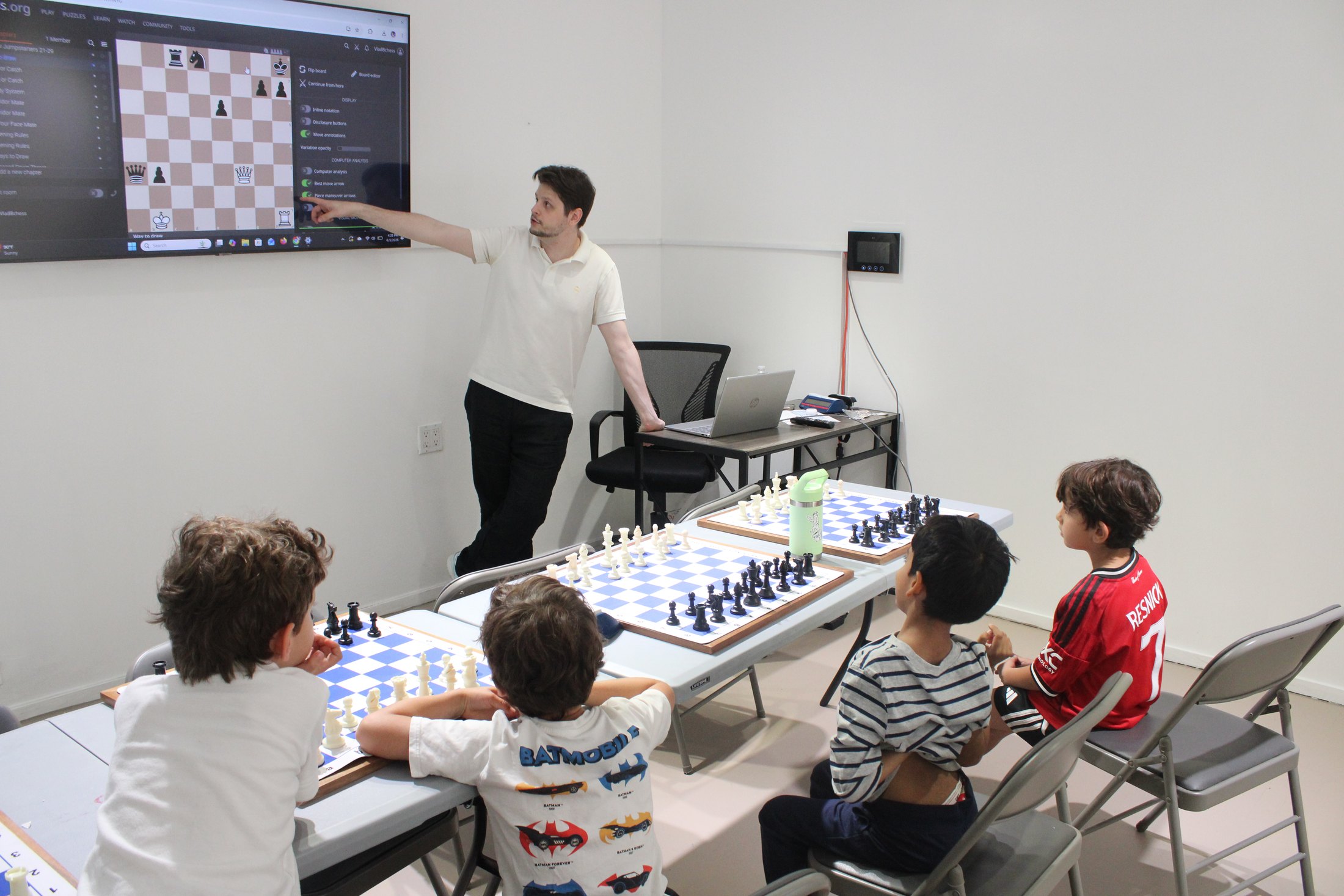 Chess coach teaching children in a group lesson with boards and a screen.